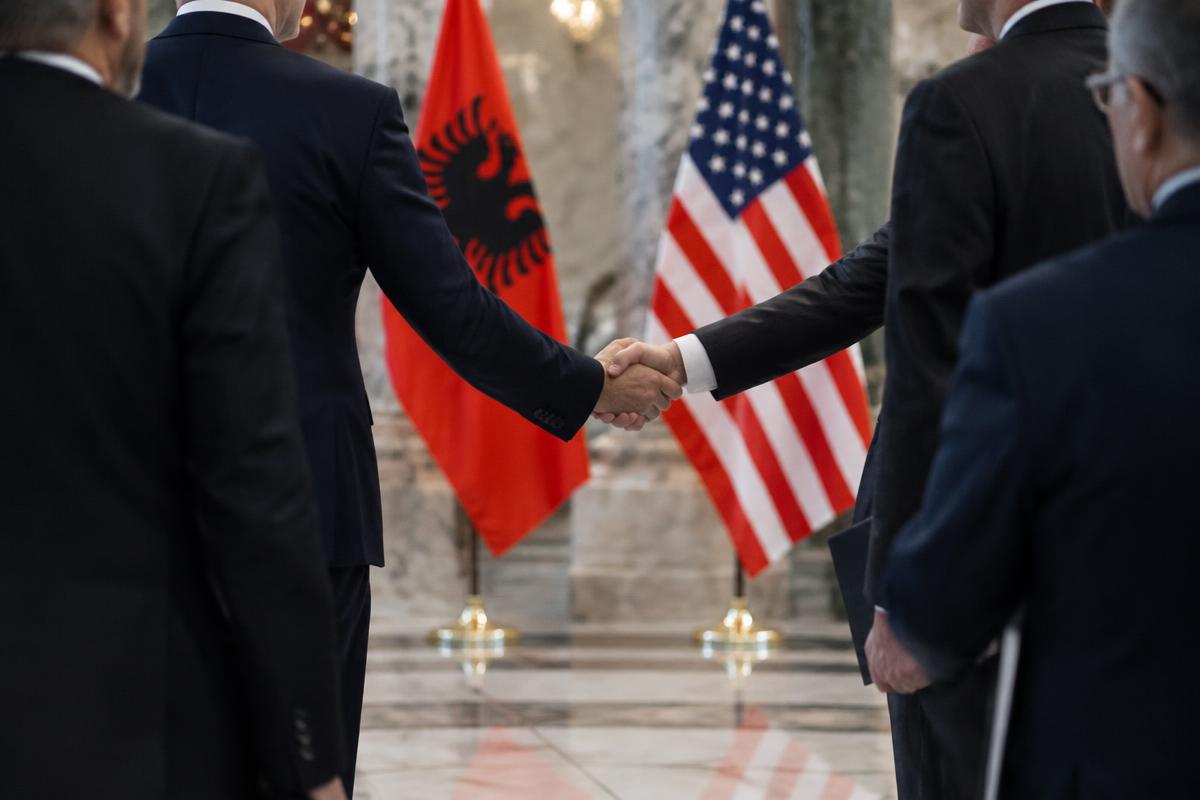 Illustration: two men in dark suits shake hands in a marble hall, with the Albanian and American flags standing behind them.