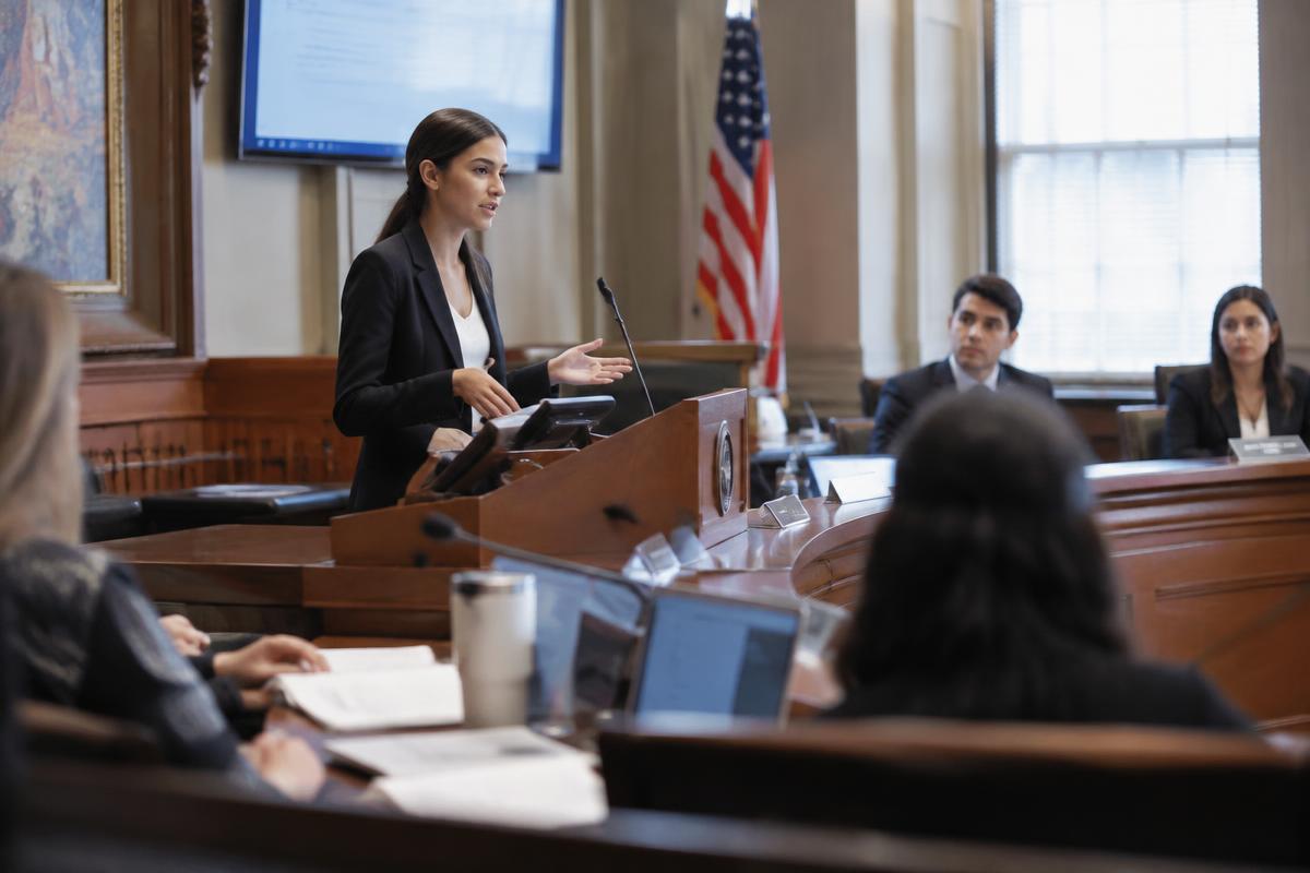 Illustration: a young woman speaks from a wooden podium in a hearing room, with the American flag standing behind her and listeners seated at the desks.